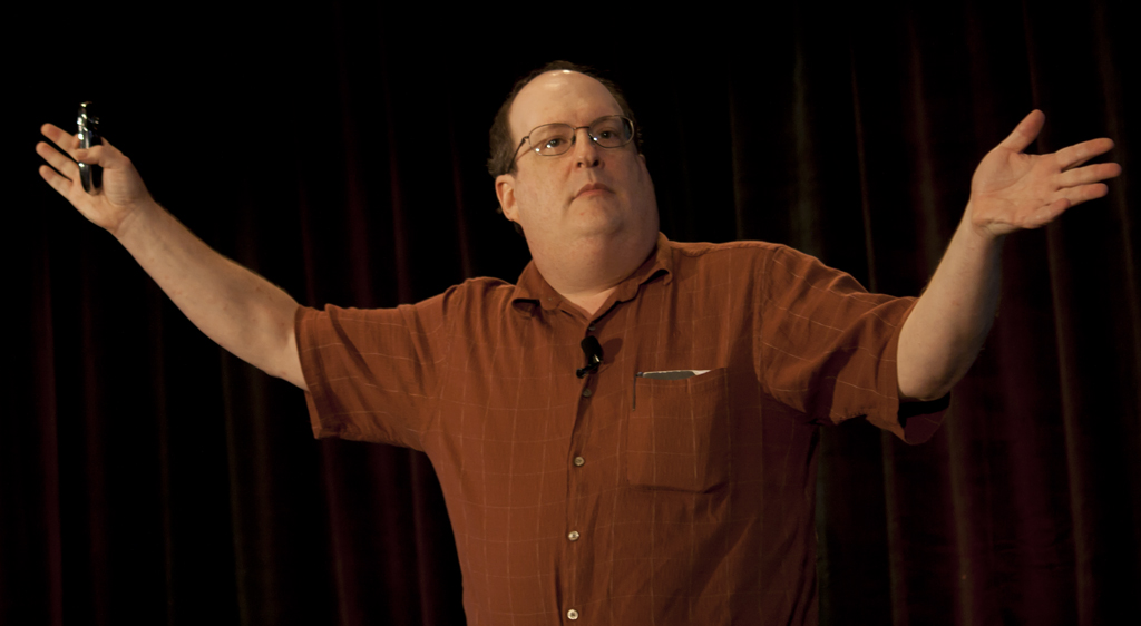 A heavy middle aged man spreads his arms wide, as if addressing a crowd